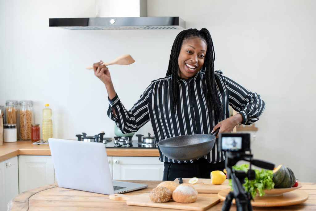 Happy african american woman vlogger broadcasting live video online teaching cooking food in kitchen at home