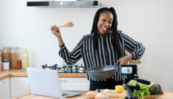 Happy african american woman vlogger broadcasting live video online teaching cooking food in kitchen at home