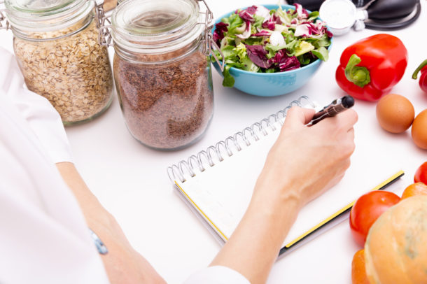 an image of a nutritionist's hand writing in a notebook, surrounded by jars of oats, a salad, eggs and red peppers