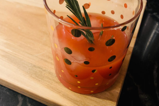 a cocktail glass filled with a red liquid and a rosemary spring sitting on a wooden cutting