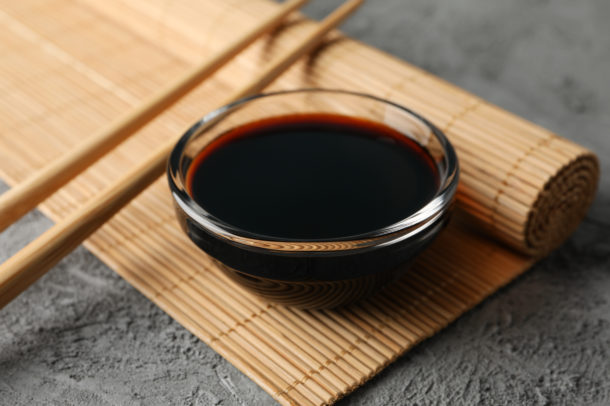 A bowl of soya sauce in a small clear bowl placed on a partially rolled bamboo placemat beside chopsticks.