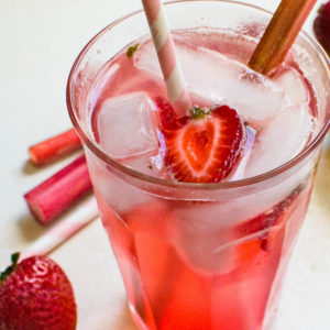 a clear glass filled wit pink lemonade with ice cubes and strawberries floating along side a straw and stalk of rhubarb