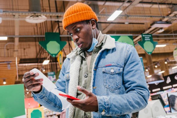 Disappointed African-American customer in denim jacket with mask looks at receipt total holding mobile phone in supermarket