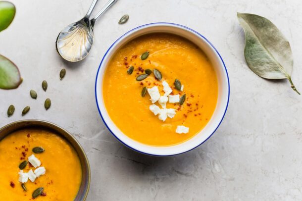 “Bowl of creamy carrot and ginger soup topped with crumbled cheese, pumpkin seeds, and a sprinkle of paprika, served on a light stone surface with spoons and scattered seeds nearby.”