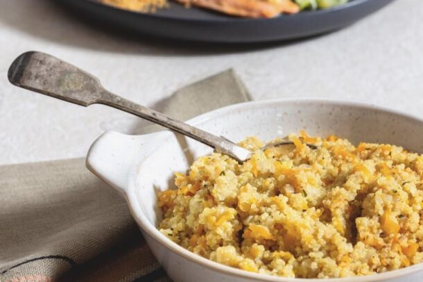 A bowl of cooked barley and grated carrots sits on a beige cloth napkin with a spoon resting inside. In the background, a plate with salmon, lemon wedges, and a vegetable garnish is slightly out of focus.