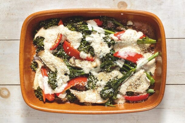 A rectangular brown baking dish filled with roasted broccolini, red bell pepper slices, and eggplant pieces drizzled with a creamy white tahini sauce and sprinkled with sesame seeds, set on a light wooden surface.
