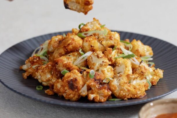 A plate of golden-brown baked cauliflower florets coated in a sweet and spicy sauce, garnished with sliced green onions. A hand holds a small fork with one piece lifted above the dish, and a bowl of dipping sauce sits nearby.