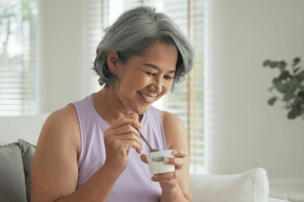 Smiling older woman with short gray hair enjoying a cup of yogurt while sitting on a couch in a bright, sunlit room.