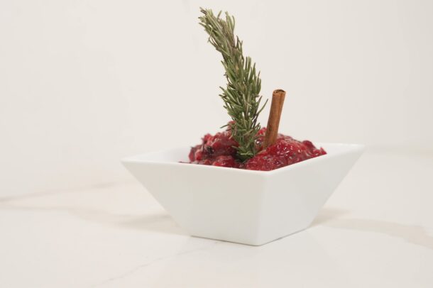 Cranberry sauce in a white square bowl garnished with a sprig of rosemary and a cinnamon stick.