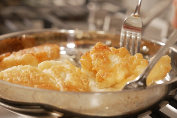 Golden, crispy bannock being lifted with a fork and spoon from a stainless steel pan on a stovetop.