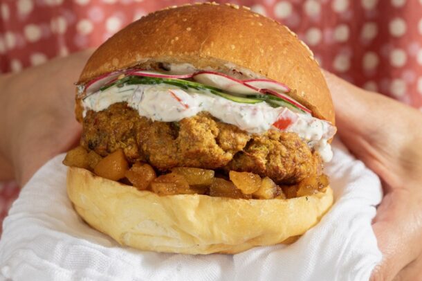 A person holds a sandwich filled with crispy fried chicken, chunky apple chutney, and creamy cucumber sauce on a soft bun against a pink patterned background.