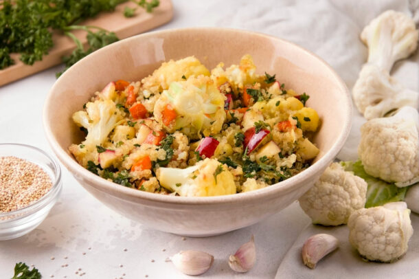 bol de quinoa avec chou-fleur rôti, carottes et fines herbes fraîches, entourée de fleurons de chou-fleur et de gousses d’ail.