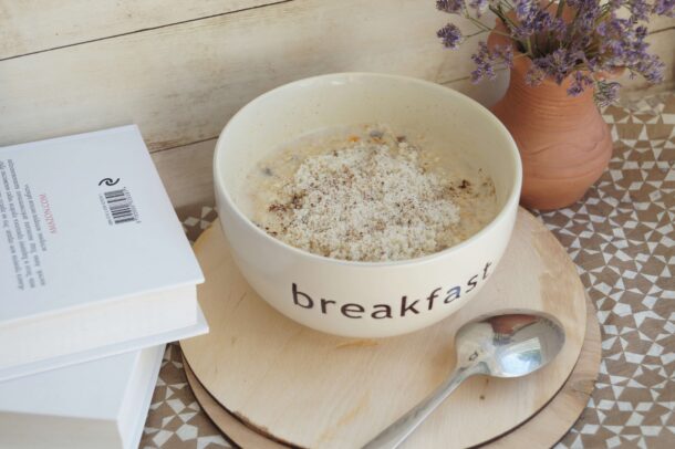 : Bowl of creamy overnight oats topped with cinnamon, served beside a spoon and a book on a breakfast table.