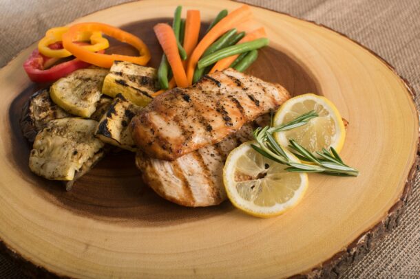 Grilled chicken breast served with roasted vegetables, green beans, carrot sticks, lemon slices, and rosemary on a wooden platter.
