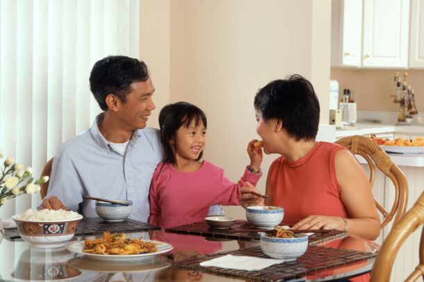 Trois personnes sont assises autour d’une table à manger, partageant un repas dans une cuisine lumineuse. Des bols, du riz et différents plats sont disposés sur la table.